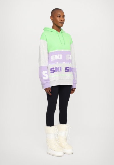Color-blocked hoodie in green, gray, and lavender with "SKI" text. Paired with black leggings and white fur-lined boots. Smooth texture.