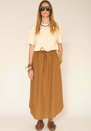 Femme portant de grandes lunettes de soleil, un haut beige à manches courtes, une jupe taille haute marron, plusieurs bracelets, et des mocassins marron, debout devant un fond uni.