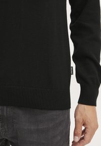Schwarzer Strickpullover mit geripptem Saum und Bündchen, aus einem weichen Material gefertigt, kombiniert mit dunkelgrauen Jeans und einem Etikett, das den Markennamen zeigt.