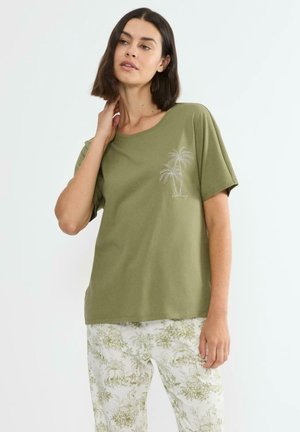 Kobieta w oliwkowej koszulce z nadrukiem palmy i jasnych kwiecistych spodniach, stojąca z ręką dotykającą szyi.