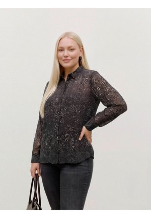 Une femme blonde porte un chemisier noir à motifs et un jean foncé, et tient un sac à main devant un fond de studio uni.