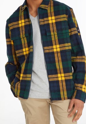 Homme portant une veste zippée à carreaux jaune, vert et marine sur une chemise gris clair et un pantalon beige, la main reposant sur le côté.