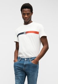 Νέος άντρας που φοράει λευκό μπλουζάκι Tommy Jeans με μπλε και κόκκινες ρίγες, συνδυασμένο με μπλε τζιν τζιν, τα χέρια στις τσέπες.