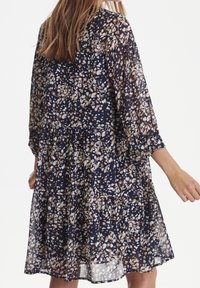 Marinblå blommig klänning i lätt och genomskinligt tyg. Har lång ärmar, lagerdesign och små krämfärgade och bruna blommönster över hela.