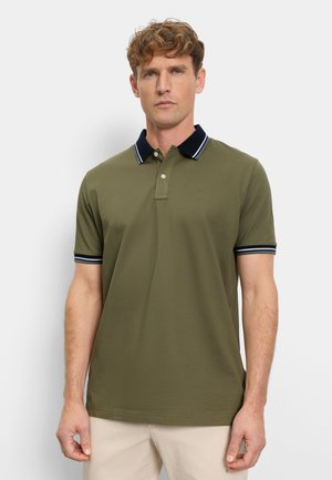 Mann trägt ein olivgrünes Poloshirt mit marineblauem Kragen und Ärmelsaum, steht vor einem einfarbigen hellen Hintergrund.