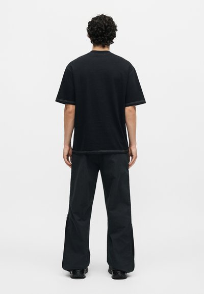 Άνδρας με σγουρά μαλλιά, στέκεται πλάτη, φοράει απλό μαύρο κοντομάνικο μπλουζάκι, φαρδιά μαύρη παντελόνια και μαύρα παπούτσια.