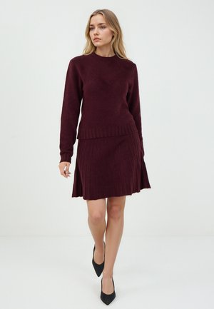 Pull en maille bordeaux à manches longues et ourlet côtelé, associé à une jupe plissée assortie. Tissu texturé et silhouette ajustée. Talons noirs.