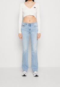 Donna che indossa un cardigan corto a coste bianco, jeans dritti azzurri e sneaker bianche, in piedi contro uno sfondo semplice.