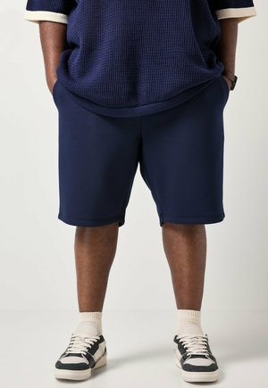 Person iført marineblå shorts, en struktureret marineblå skjorte med cremefarvede manchetter, hvide sokker, sort-hvide sneakers, hænderne i lommerne.