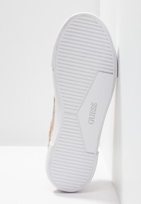 Podešev tenisky s bílou pryží s drážkami, horní část ve zlaté textuře. Logo "GUESS" na boku, minimalistický design, lehká konstrukce.