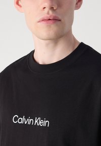Svart bomull T-skjorte med rund hals og en hvit "Calvin Klein" logo på forsiden. Stoffet er glatt med minimal tekstur.