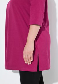 Fuchsia-Tunika-Oberteil mit lockerer Passform, dreiviertel Ärmeln, seitlichen Schlitzdetails und weichem, leichtem Baumwollstoff. Kombiniert mit schwarzen Leggings.