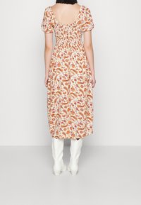 Floral midi-jurk in licht perzik met een paisleypatroon, korte puffmouwen en een smock achter. Gecombineerd met kniehoge witte laarzen.