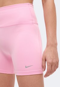 Άτομο που φοράει ροζ αθλητικά σορτς Nike με γκρι λογότυπο Swoosh στον αριστερό μηρό και ψηλή μέση.