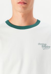 Camiseta blanca con cuello acanalado verde, que presenta el logo "Cowboy Country" en verde claro en el lado izquierdo, hecha de tela de algodón.