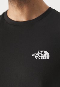 Detailní záběr osoby v černém tričku s bílým logem The North Face na hrudi poblíž výstřihu.