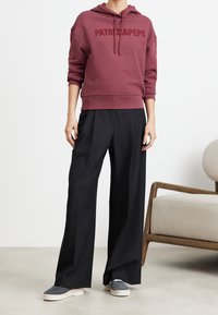 Felpa con cappuccio color mogano con logo "PATRIZAPPEPE", abbinata a pantaloni larghi neri e scarpe slip-on grigie. Design minimalista, look casual.