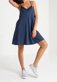 Laivansinis hihaton mekko, jossa on pitsireunus, A-linjainen siluetti, teksturoitu kangas, yhdistetty harmaisiin tennareihin, seisoo yhdellä jalalla.