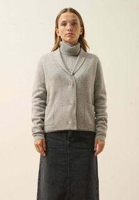 Cardigan gris con cuello de chal, cierre de botones y dos bolsillos frontales, llevado sobre un suéter de cuello alto gris claro y una falda de mezclilla. Textura suave.