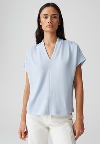 Blouse bleu clair à manches courtes avec un col en V, en tissu texturé. Présente une coupe ample et un léger plissé aux épaules.