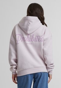 Haut à capuche rose clair avec une capuche spacieuse, des poignets côtelés et un graphique "Interdit" au dos dans une police cursive audacieuse. Matériau en mélange de coton.