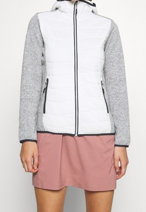 Femme portant une veste matelassée blanche avec des manches en maille grises, une fermeture éclair à l'avant et une jupe rose sur un fond clair uni.