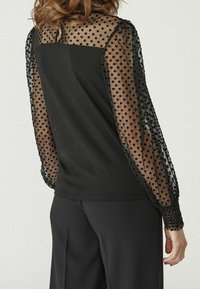 Haut noir avec manches transparentes à pois et poignets élastiques. Le corps est entièrement noir, contrastant avec les manches texturées et transparentes.