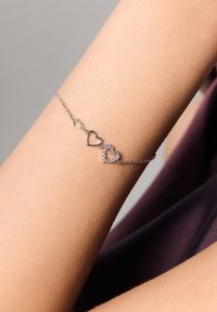 Srebrna bransoletka z trzema zawieszkami w kształcie serc, z czego dwa są puste, a jeden ozdobiony przezroczystymi kamieniami, noszona na nagim nadgarstku.