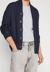 Námořnický pletený kabátek s kabelovou texturou, se šesti hnědými knoflíky a šálovým límcem, nošený přes šedé tričko a světlé šedé džíny.