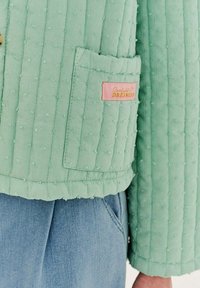 Prošívaná bunda mátové barvy s texturovaným povrchem, vybavená vertikálními švy a náprsní kapsou. Obsahuje růžovou etiketu "Sunlight Dreamers".