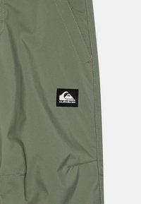 Gröna vattentäta byxor med ett Quiksilver-logotypmärke. Slät textur med synliga sömmar och en sidofickdetalj.