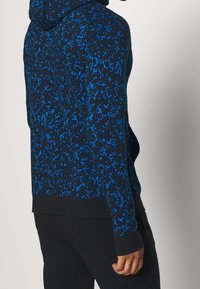 Sweat à capuche avec un motif éclaboussé bleu et noir, fabriqué en tissu doux. Dispose d'un ourlet et de poignets côtelés. Capuche avec cordon de serrage réglable.