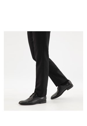 Person trägt schwarze formelle Schuhe und schwarze Hosen und geht auf einem weißen Hintergrund, rechter Fuß in der Luft, mittlerer Schritt.