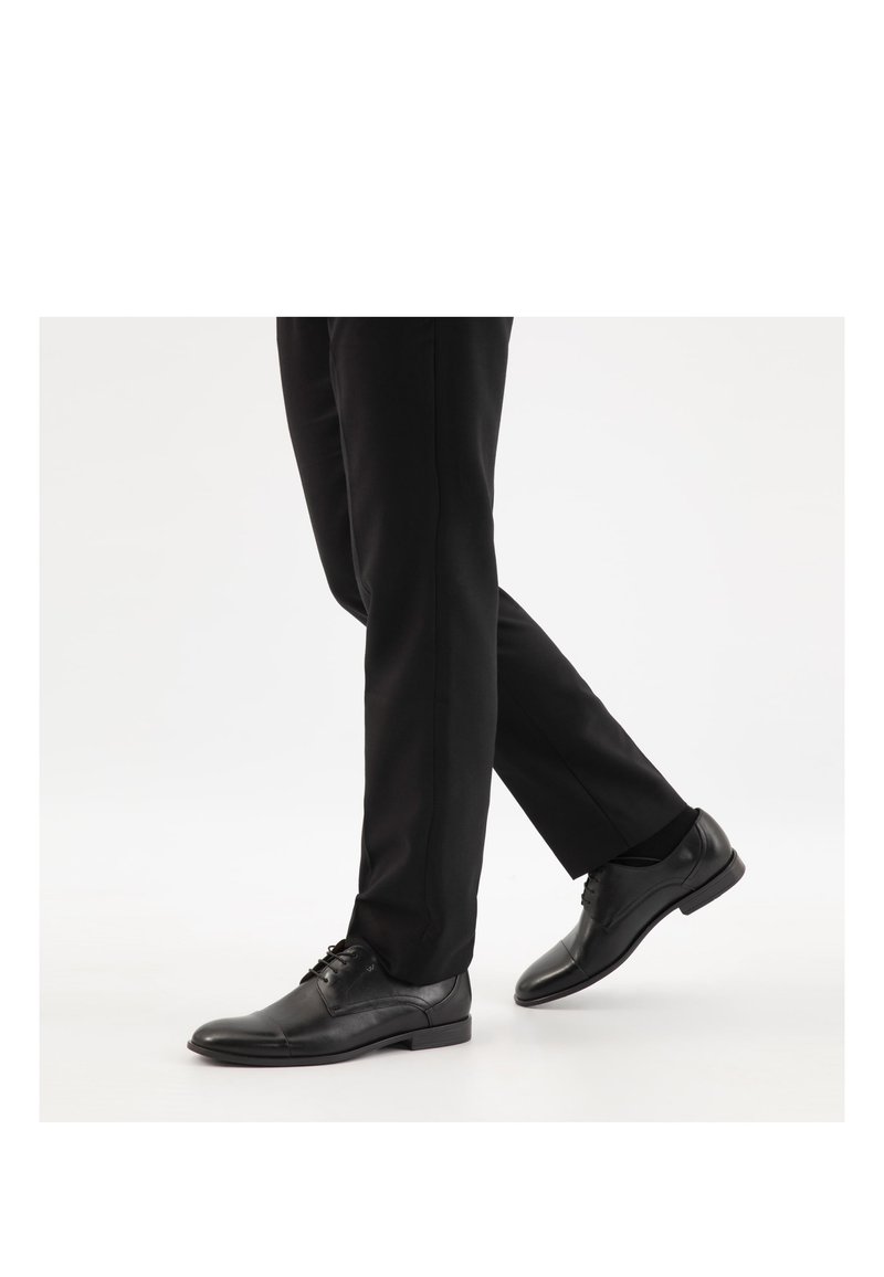 Persoon draagt zwarte nette schoenen en zwarte broek, wandelend op een witte achtergrond, met de rechtervoet halverwege een stap in de lucht.