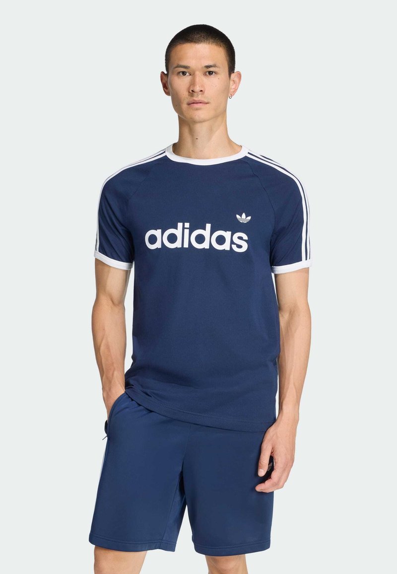 Tmavomodré tričko Adidas s bielym logom a pruhmi, krátkymi rukávmi a okrúhlym výstrihom, doplnené o zodpovedajúce tmavomodré šortky.