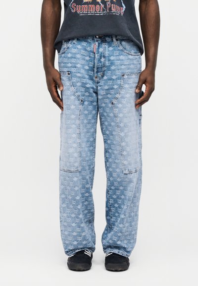 Φαρδιά τζιν σε ανοιχτό γαλάζιο με τσέπες-μπαλώματα και διακριτικό τετράγωνο μοτίβο σε όλο το ύφασμα, φορεμένα με μαύρα αθλητικά παπούτσια και σκούρο graphic t-shirt.