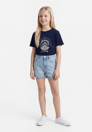 Chica rubia sonriendo, vistiendo una camiseta azul marino con un gráfico de concha y perla, pantalones cortos de mezclilla clara y zapatillas blancas, de pie sobre fondo blanco.