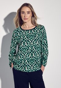 Grön och vit mönstrad blus med rund halsringning, långa ärmar med samlad detalj och slät textur. Matchas med mörka byxor.