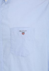 Jasnoniebieska koszula na guziki z naszywką z logo „GANT” oraz małą haftowaną tarczą w kolorach czerwonym, białym i niebieskim.