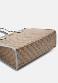 Bolso tote de color beige con un patrón y detalles en cuero azul. Presenta acentos dorados y tachuelas, con un fondo estructurado y dos asas para llevarlo.