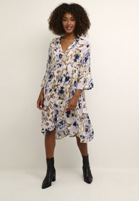 Robe florale dans des couleurs claires avec des motifs bleus et beige, décolleté en V, manches cloche trois-quarts et ourlet asymétrique fluide. Bottines noires.