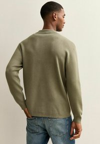 Olivgrüner Strickpullover mit langen Ärmeln, einfachem Kragen und lockerer Passform. Kombiniert mit blauen Jeans aus Denim, die Gesäßtaschen haben.