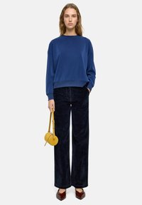 Niebieski, oversize'owy sweatshirt z ryflowanymi mankietami, połączony z ciemnymi spodniami dzwonami. Akcesoria to mała żółta torebka i ciemno-brązowe, spiczaste buty.