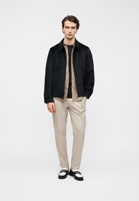 Giacca in suede nera sopra una camicia beige chiaro e una t-shirt scura, abbinata a pantaloni leggeri a quadri e mocassini bicolore. Design semplice e pulito.