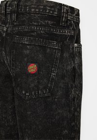 Calças de ganga pretas com uma textura desbotada, bolso traseiro com um patch de logotipo "Santa Cruz" em vermelho e amarelo, e detalhes de costura padrão.