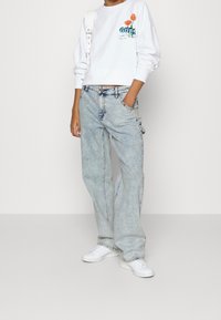 Vit sweatshirt med orange blomgrafik och "Guess"-logotyp, tillsammans med ljusblå baggyjeans och vita sneakers.