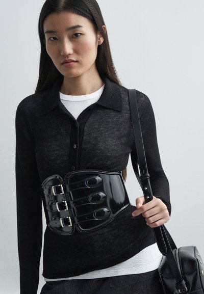 Zwarte lakleren tailleband met vier verstelbare gespen, gedragen over een zwarte transparante top en een witte coltrui, met een schoudertas.