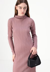 Ribota mezgimo turtleneck suknelė švelnios mauve spalvos, derinama su juodu odiniu rankiniu krepšiu, turinčiu kreivinę rankeną ir logotipo akcentą.