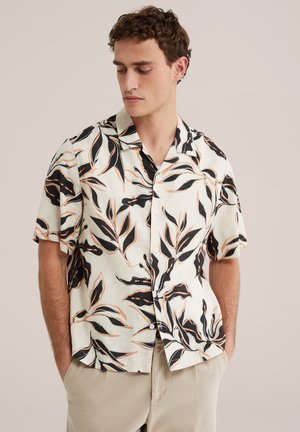 Muž nosiaci košeľu s krátkym rukávom s veľkými čiernymi a hnedými listovými vzormi, ruky v kapsách béžových nohavíc, pozerá dole.