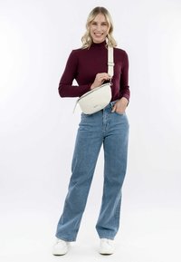 Sac en bandoulière beige avec une finition texturée et une fermeture éclair, porté par une personne vêtue d'un haut ajusté bordeaux et d'un jean taille haute bleu.